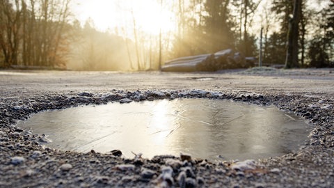 Die Sonne scheint an einem Wanderparkplatz auf das gefrorene Wasser eines Schlaglochs.