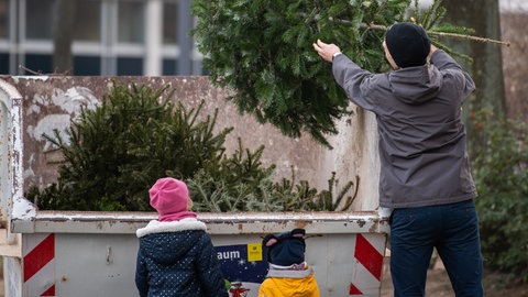 Eine Person steht mit zwei Kindern vor einem großen Container, in den er seinen ausgedienten Weihnachtsbaum wirft. Die Bäume werden werden oft zu Kompost oder Holzhackschnitzeln verarbeitet, um einen Beitrag zur regionalen Ressourcennutzung zu leisten. 