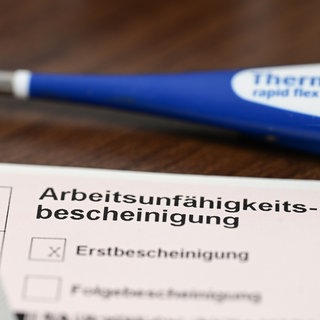 Ein Fieberthermometer, Medikamente und eine ärztliche Arbeitsunfähigkeitsbescheinigung (Krankmeldung) liegen auf einem Nachttisch.