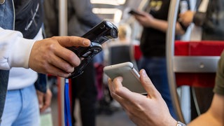 Symbolbild | Ein Fahrkartenkontrolleur (l) kontrolliert mit einem elektronischen Lesegerät den elektronischen Fahrschein eines Fahrgasts.