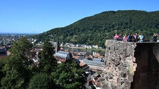 Touristen stehen am Heidelberger Schloss bei strahlendem Sonnenschein an einem Aussichtspunkt und schauen und fotografieren in die Altstadt.