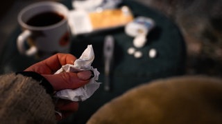 Eine Frau hält ein Taschentuch in ihrer Hand. Im Hintergrund steht eine Tasse Tee und ein Fiebertherometer und Medikamente liegen auf einem Tisch. 