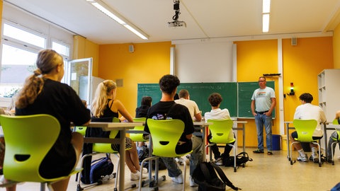 Schülerinnen und Schüler sitzen in einem Klassenzimmer auf grünen Stühlen. Vor einer Tafel steht ein Lehrer.