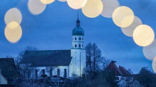 Blick am Morgen, kurz vor Sonnenaufgang durch eine Lichterkette auf die beleuchtete Pfarrkirche Mariä Himmelfahrt von Seekirch am Federsee (Kreis Biberach).