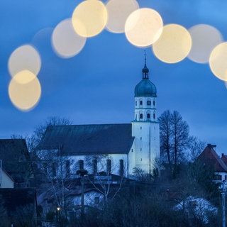 Blick am Morgen, kurz vor Sonnenaufgang durch eine Lichterkette auf die beleuchtete Pfarrkirche Mariä Himmelfahrt von Seekirch am Federsee (Kreis Biberach).