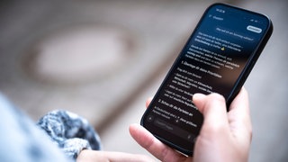 Eine Person hält ein Smartphone in der Hand und liest in der Anwendung ChatGPT eine Antwort auf die Frage «Was soll ich am Sonntag wählen?». 