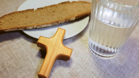 Ein Glas Wasser, ein Kreuz aus Holz, und eine Scheibe Brot auf einem Teller. An Karfreitag verzichten viele Menschen auf Fleisch. Doch es gibt auch moderne Formen des Fastens.