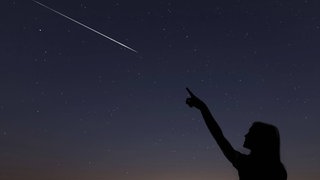 Frau zeigt auf Sternschnuppe am Nachthimmel. Die Lyriden sind bald am Himmel zu sehen. In der Nacht vom 22. auf 23. April erreicht der Sternschnuppenschauer sein Maximum.
