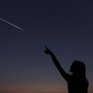 Frau zeigt auf Sternschnuppe am Nachthimmel. Die Lyriden sind bald am Himmel zu sehen. In der Nacht vom 22. auf 23. April erreicht der Sternschnuppenschauer sein Maximum.