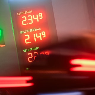 Ein verschwommen zu sehenes Auto fährt an einer Tankstelle dabei. Dort sind hohe Preise für Diesel und Benzin zu sehen.