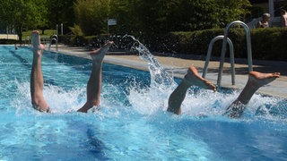 Junge Männer springen im Freibad ins Wasser.