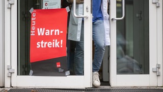 Ein Plakat mit der Aufschrift "Heute Warnstreik" hängt am Eingang des Gebäudes eines Universitätsklinikums.