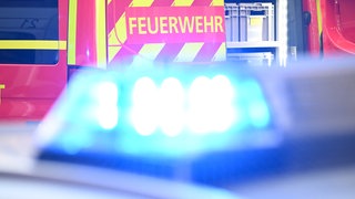 Ein Fahrzeug der Feuerwehr steht bei einem Einsatz auf einer Straße, während im Vordergrund das Blaulicht eines Polizeifahrzeugs zu sehen ist.