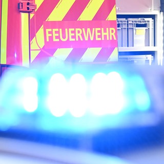 Ein Fahrzeug der Feuerwehr steht bei einem Einsatz auf einer Straße, während im Vordergrund das Blaulicht eines Polizeifahrzeugs zu sehen ist.