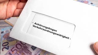 Ein Brief mit der Aufschrift "Anhörungsbogen Verkehrsordnungswidrigkeit" wird vor dem Hintergrund von Euroscheinen in der Hand gehalten.