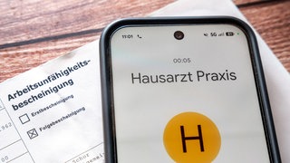 Ein Smartphone mit Anruf beim Hausarzt liegt neben einer Arbeitsunfähigkeitsbescheinigung: Die Bundesregierung plant eine Krankschreibung ab dem ersten Tag (neue Reform) und das Ende der telefonischen Krankschreibung.