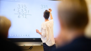 Eine Referendarin schreibt eine Mathematikaufgabe auf eine digitale Schultafel in einem Klassenraum. 