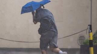 Ein Mann rennt in Stuttgart durch starken Regen und versucht, sich mit einem Regenschirm zu schützen. Ein Blitzeinschlag am Hauptbahnhof sorgte für Verspätungen bei der Bahn.