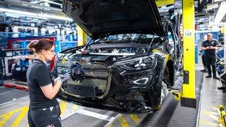 Beschäftigte arbeiten in einer Halle in einem Mercedes-Benz Werk an einem Fließband an Fahrzeugen vom Typ GLC.