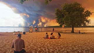 Strandbesuchende sitzen am Strand von Moutchic in Lacanau. Im Hintergrund sind Rauchwolken von den Waldbränden.