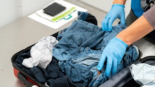 Bei einer Zollkontrolle am Flughafen öffnet ein Beamter sorgfältig das Gepäck eines Reisenden. Der Koffer wird auf verdächtige Gegenstände geprüft.