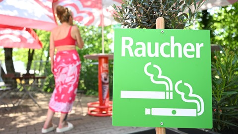Eine Frau steht rauchend in einem ausgewiesenen Raucherbereich in einem Freibad neben einem grünen Hinweisschild mit der Aufschrift "Raucher".