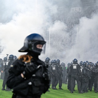 Ein Polizist steht beim Fußballspiel zwischen Waldhof Mannheim und dem 1. FC Kaiserslautern auf dem Platz. Im Hintergrund stehen zahlreiche weitere schwarz gekleidete Polizisten auf dem Rasen. Hinter ihnen steigt Rauch auf.