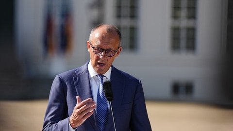Bundeskanzler Friedrich Merz (CDU) in blauem Anzug mit blau-gepunkteter Krawatte und Brille vor einem Mikrofon.