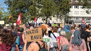 Viele Menschen stehen auf dem Platz an der Alten Synagoge in Freiburg. Dort findet eine Demonstration gegen Rechtsextremismus statt. Eine Teilnehmeriin hält ein Schild, auf dem "Nazis raus" steht.