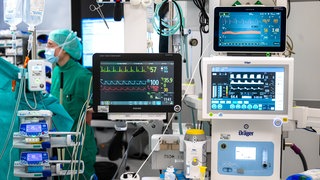 In einem Operationssaal eines Krankenhauses sind mehrere Monitore zu sehen, die Vitaldaten anzeigen. Im Hintergrund sind Ärzte zu sehen. (Symbolfoto)