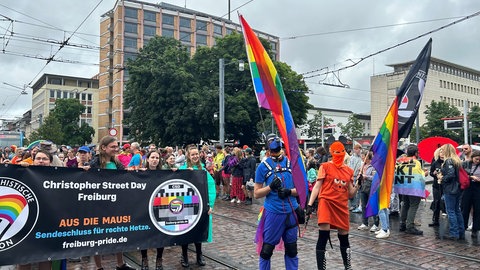 Mehrere Leute halten einen Banner - davor stehen zwei vermummte Leute in den Farben von Maus und Elefant aus der Sesamstraße.