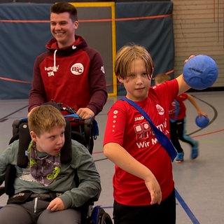 Ein Kind wirft einen Ball. Ein Kind im Rollstuhl sieht ihm dabei zu.