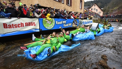 Auf einem Bach fahren selbstgebaute Boote. Auf ihnen sitzt eine Band, die ihre Instrumente spielt. Neben dem Bach jubeln Menschen ihnen zu.