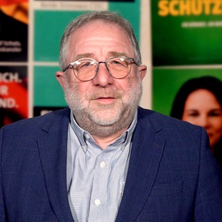 Mann in blauem Jackett steht vor einer Wand aus mehreren Wahlplakaten. Der Kommentator hat eine Brille und graue Haare. 
