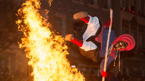 Eine Kandelhexe mit einem Stock in der Hand springt über eine große Flamme
