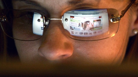 In der Brille einer jungen Frau spiegelt sich eine Internetseite zur Partnersuche.