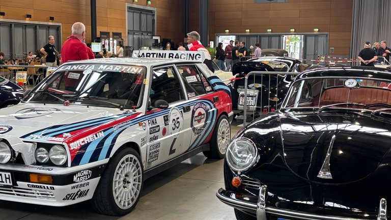 Zwei Oldtimer stehen nebeneinander, ein klassisches schwarzes und ein neuerer, weißer Sportwagen.