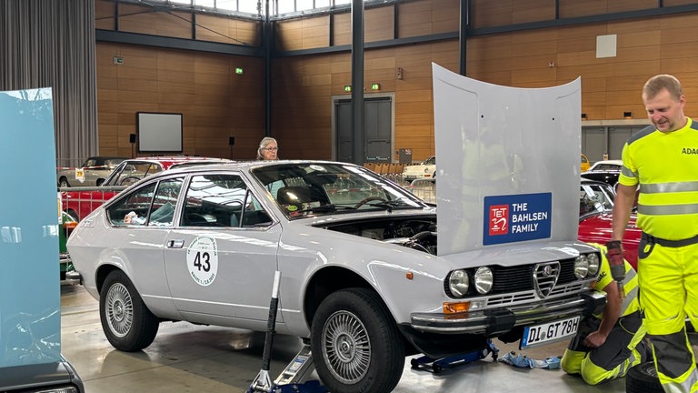 Ein graues Oldtimer-Ato steht auf einem Wagenheber. Mehre Männer mit neongelben Anzügen hocken und stehen daneben.