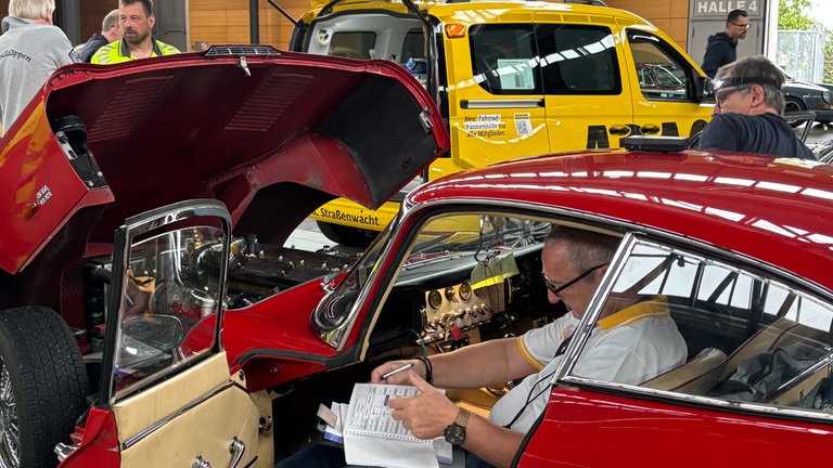 Ein Mann sitzt in einem roten Oldtimer-Auto und blättert in einer Broschüre.