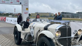 Ein weißer Oldtimer, Baujahr 1930, fährt durch eine Startbahn. Ein Mann und eine Frau sitzen mit lederner Fliegermütze in dem Fahrzeug.