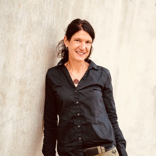 Frau mit langen schwarzen Haaren lehnt an einer Wand. Sie trägt eine dunkle Bluse und lächelt in die Kamera.