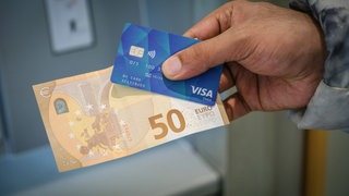Ein Mann hält eine Bezahlkarte für Asylsuchende und einem 50 Euro Schein in der Hand.