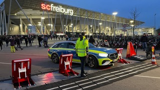 Ein Polizist steht neben einem Polizeiauto an einer Fahrzeugsperre vor dem Europa Park-Stadion in Freiburg 