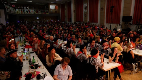 Verkleidete Personen sitzen an Tischen in einer Halle und klatschen.