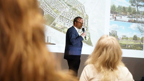 Ein Mann steht auf einer Bühne. Hinter ihm wird eine Leinwand mit einem Bild des neuen Stadtteils Dietenbach angestrahlt.
