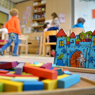 In einer Kindertagesstätte liegt Spielzeug auf dem Boden. Im Hintergrund spilen Kinder betreut durch eine Erzieherin. 