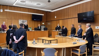 Fast zweieinhalb Jahre nach einem Mord an einer Offenburger Schule müssen sich die Eltern des damals 15-jährigen Täters wegen fahrlässiger Tötung vor Gericht verantworten. Sie stehen und sitzen auf der rechten Seite mit ihren Verteidigern. Ihnen gegenüber stehen die Eltern des gleichaltrigen Mitschülers gegenüber, der mitten im Klassenzimmer erschossen wurde.