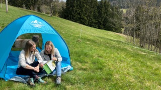 Zwei Frauen sitzen vor einem Zelt in der Natur.