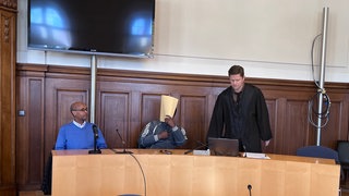Der Angeklagte sitzt zwischen seinem Dolmetscher und seinem Verteidiger auf der Anklagebank