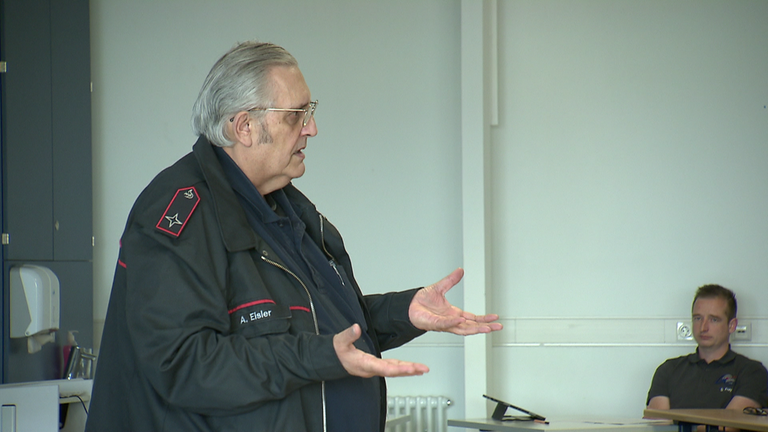 Ein in Feuerwehr uniformierter Mann steht mit ausgestreckten Armen in einem Klassenzimmer und redet.
